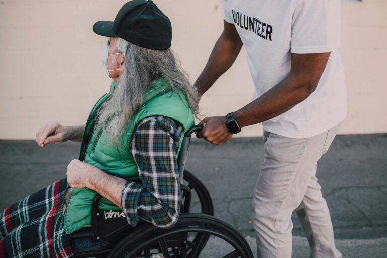 A volunteer helps a senior in a wheelchair during daytime outdoors.